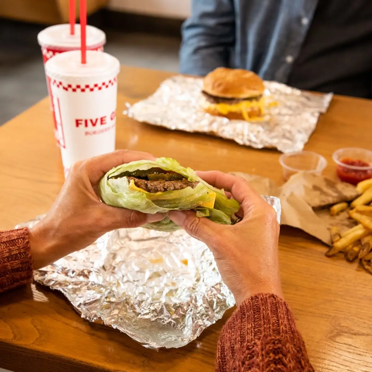 Customer's Hands Hold a Five Guys Lettuce Wrap at a Dining Room Table Inside a Five Guys Restaurant